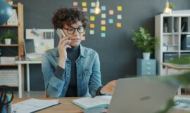 Woman talking on phone at desk with laptop.